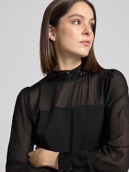 Kobiece model prezentuje bluzka z długim rękawem z cekinowym obszyciem od Apricot, elastyczna mieszanka materiałów, krój regular fit, stójka, półprzezroczysty materiał, bufiaste rękawy, długość tyłu w rozmiarze S: 58 cm, czarny kolor, tekstura matowa, detaliczne ujęcie górnej części ciała.