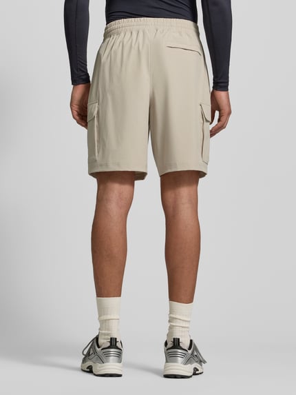 Mannelijk model draagt Straight Fit Shorts met cargozakken model 'Vibe' van Under Armour in beige, elastische tailleband, logo, rechte fit, cargo- en achterzak, materiaal mix, hele lichaamsopname achteraan.