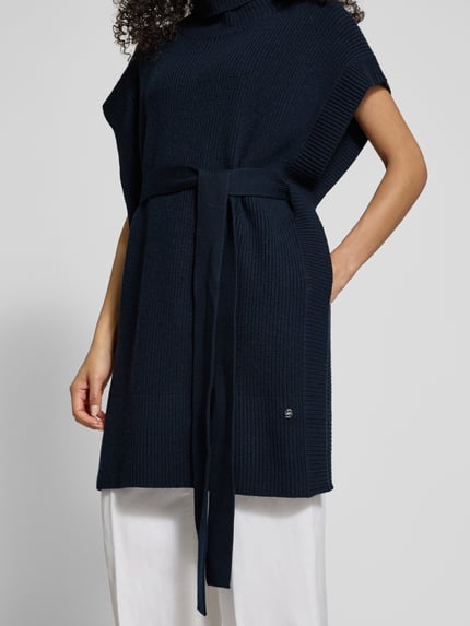 Frauenmodel trägt Regular Fit Poncho aus reiner Schurwolle Modell 'FAVANDA' von BOSS, Optik: Unifarben, Strickoptik, mit Bindegürtel, Logo-Applikation, Rollkragen, Ärmelform: Ohne, Materialverarbeitung: Strick, Ganzkörperansicht von vorne.
