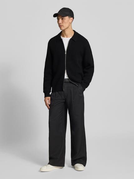Männermodel trägt Cardigan aus reiner Wolle von Filippa K in Schwarz mit Ripp-Optik und Zweiwege-Reißverschluss, Ganzkörperansicht von vorne.