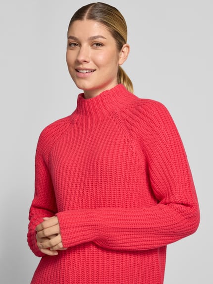 Frauenmodel trägt Strickpullover aus Kaschmir-Baumwoll-Mix Modell 'LONA' von 0039 Italy in leuchtendem Pink, grobe Strickstruktur, Nahaufnahme Oberkörper. Rückenlänge 60 cm, Ärmellänge 78 cm, Regular Fit, Stehkragen.