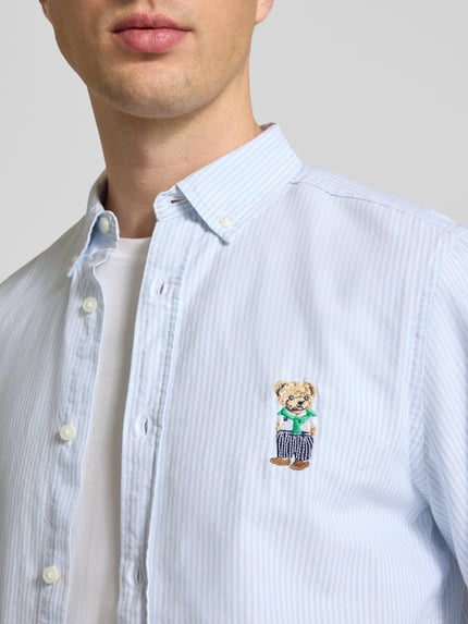 Męski model nosi Slim Fit Freizeithemd mit Button-Down-Kragen von CHILL PILL z bawełny, wyhaftowany motyw, jasnoniebieskie paski, Nahaufnahme górnej części ciała. Długość tyłu w rozmiarze M: 79 cm, Szerokość w rozmiarze M: 44 cm, Długość rękawów w rozmiarze M: 67 cm.