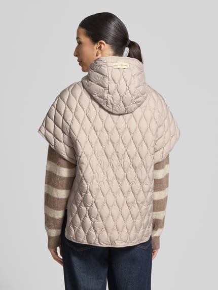 Modelka nosi Steppjacke im kurzärmeligen Design od Fuchs Schmitt, luźny krój, kaptur, zamek, krótkie rękawy, kieszenie, beż, widok całej sylwetki z tyłu, długość tyłu 65 cm, szerokość ramion 47 cm, szerokość w rozmiarze 36: 57 cm.