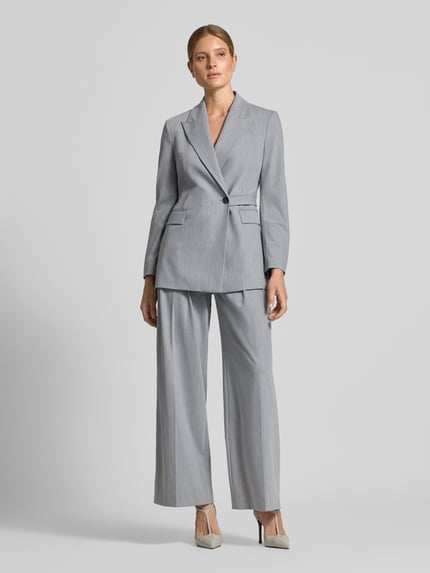 Frauenmodel trägt Fließende Anzughose in Mittelgrau Melange - curated by Viky Rader von Style Icon Collection, Relaxed Fit, Wide Leg, Kellerfalten, Knopf- und Reißverschluss, strukturierte Stofftextur, Ganzkörperansicht von vorne.