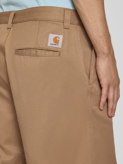 Männermodel trägt Bermudas mit Baumwoll-Anteil Modell 'Brader' von Carhartt Work In Progress in Beige, Eingriff- und Gesäßtaschen, Nahaufnahme Oberkörper. Material-Mix, Relaxed Fit, Knopf- und Reißverschluss, Gürtelschlaufen, Bundweite 44 cm, Innenbeinlänge 30 cm.