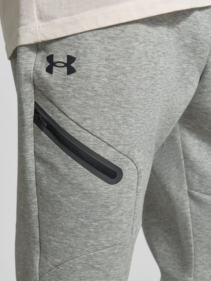 Männermodel trägt Regular Fit Sweatpants mit Logo-Print Modell 'Unstoppable' van Under Armour, elastische materiaalmix met katoen, zak op de pijp met ritssluiting, elastische band, elastische pijpboorden, binnenbeenlengte 73 cm bij maat M, detailaufnahme rechterbovenbeen.