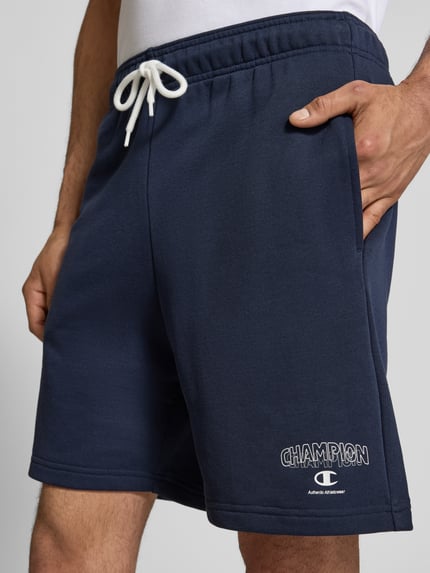Männermodel trägt Sweatshorts mit Logo und elastischem Bund von CHAMPION, dunkelblauer Baumwoll-Mix, sichtbarer Tunnelzug, Nahaufnahme Oberkörper.