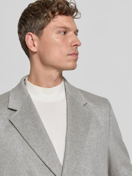 Männermodel trägt Mantel aus Wolle Kaschmir mit Westeneinsatz von BOSS in Hellgrau, Reverskragen, Strukturmuster, Nahaufnahme Oberkörper.
