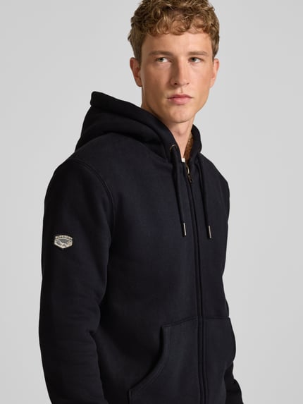 Męski model nosi Regular Fit Sweatjacke mit geteilter Känguru-Tasche Modell 'Borg' von Superdry w kolorze czarnym, kaptur z tunel, struktura miękka, długość tyłu 71 cm, długość rękawów 72 cm, bluza rozpinana, kieszeń kangurka, widok na górną część ciała z przodu.