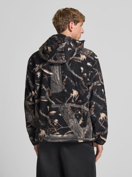 Männermodel trägt Fleecejacke mit Kapuze Modell 'Blevin' von Carhartt Work In Progress in braun-schwarzem Tarnmuster, Ganzkörperansicht Rückansicht. Gerader Schnitt, Zwei-Wege-Reißverschluss, Kapuze, Seitentaschen, Label-Patch, Rückenlänge bei Größe M 67 cm, Ärmellänge 71 cm.