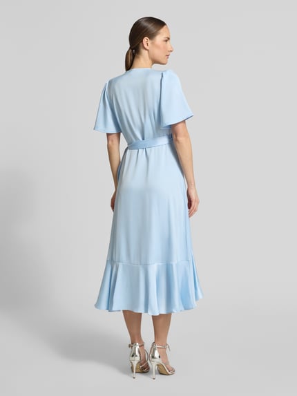 Frauenmodel trägt Cocktailkleid mit Volantsaum Modell 'THEA' von YAS aus blauem Satin, taillierter Schnitt, Midikleid, Schmetterlingsärmel, Ganzkörperansicht Rückansicht.