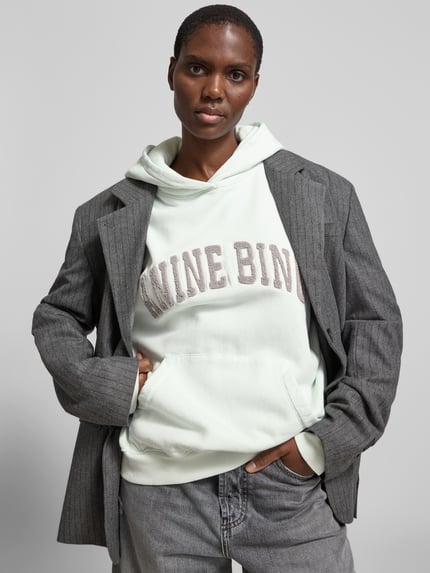 Vrouwenmodel draagt Oversized Hoodie met Logo-Stitching van Anine Bing in katoen, oversized fit, kap, kangoeroezak, voorkant borstzak, bovenlijf close-up.