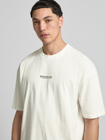 Männermodel trägt Oversized T-Shirt aus Baumwolle Modell 'Stavoren' von Pegador in Weiß, Label-Detail, Halbarm, Nahaufnahme Oberkörper. Herren T-Shirt, Reine Baumwolle, Oversized-Fit, Rundhalsausschnitt, Rückenlänge bei Größe M: 70 cm.
