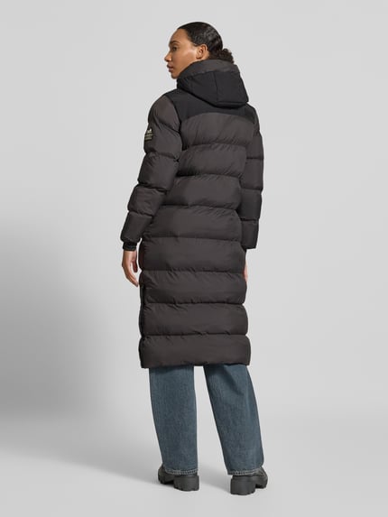 Frauenmodel trägt schwarzen Steppmantel mit Kapuze und verdecktem Reißverschluss von Superdry, strukturierte Oberfläche, Ganzkörperansicht Rückansicht. Rückenlänge 108 cm, Schulterbreite 42 cm, Brustweite 53 cm, Ärmellänge 61 cm, Regular Fit, Eingrifftaschen.