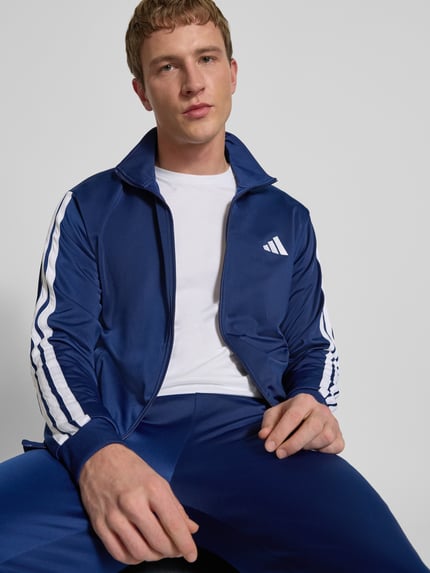 Männermodel trägt Trainingsanzug mit elastischen Abschlüssen von adidas Sportswear in Blau mit weißen Label-Details, Nahaufnahme Oberkörper.