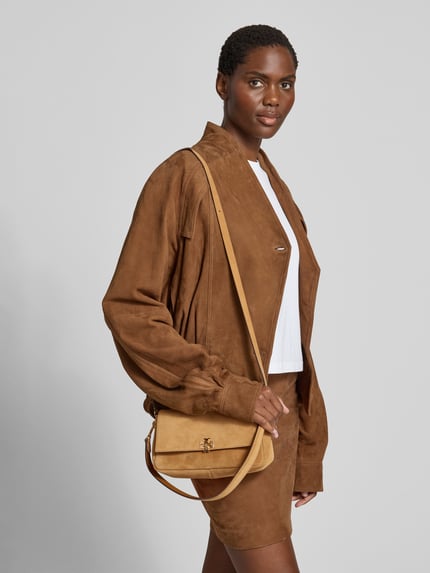 Vrouwenmodel draagt Shoulder Bag in Suede-Optik van Tory Burch in beige met logodetail, afneembare schouderband, suèdelook, hoofdvak met overslag, binnenvak met ritssluiting, breedte 24 cm, lengte 12 cm, diepte 8 cm. Ganzkörperansicht van voren.