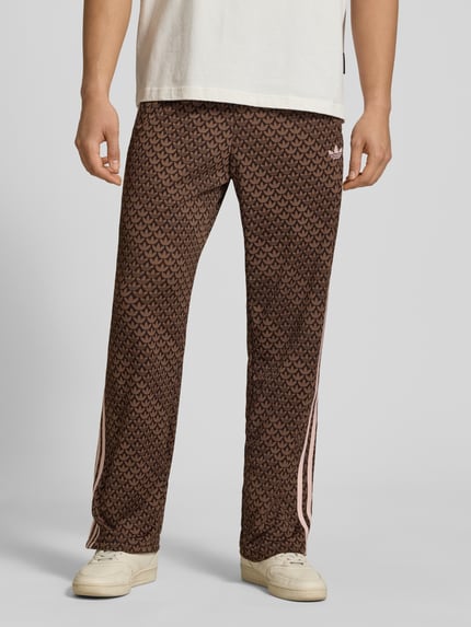 Männermodel trägt Loose Fit Sweatpants mit Logo-Muster Modell 'FIREBIRD' von adidas Originals, braune Farben, Sweat, Ganzkörperansicht von vorne.