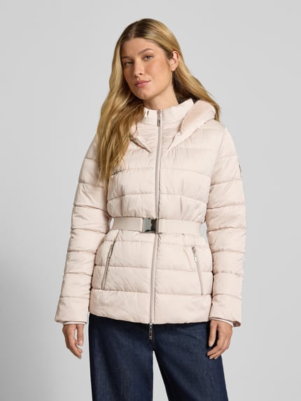 Frauenmodel trägt Steppjacke mit Reißverschlusstaschen und Gürtel von monari in hellem Beige, Steppstruktur, Zweiwege-Reißverschluss, Kunstfellkapuze, Slim Fit, Ganzkörperansicht von vorne.