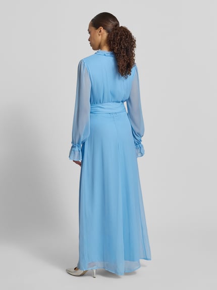 Frauenmodel trägt Maxikleid mit Gürtel und Rüschen Modell 'EMMA' von Vila aus hellblauem Chiffon in A-Linie mit elastischen Ärmelabschlüssen, Dornschließe und seitlichem Nahtreißverschluss, Ganzkörperansicht Rückansicht.