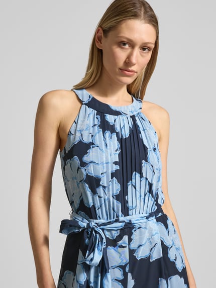 Frauenmodel trägt Jumpsuit mit Bindegürtel und Plisseefalten von Christian Berg Cocktail in Blau mit floralem Muster, Nahaufnahme Oberkörper. Taillierter Schnitt, Rundhalsausschnitt, Chiffon-Oberteil mit Plisseefalten, Bindegürtel, zarte Textur.