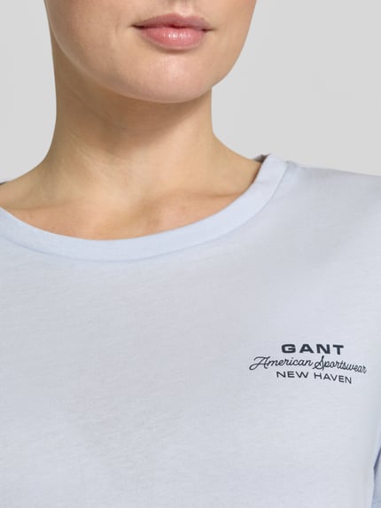 Kobieta nosi t-shirt z nadrukiem z logo i okrągłym dekoltem od Gant z czystej bawełny, krój regular fit, długość tyłu 57 cm, zbliżenie na górną część torsu.