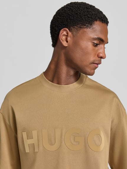 Männermodel trägt Regular Fit T-Shirt aus reiner Baumwolle Modell 'Dinkee' von HUGO in Beige mit großem Schriftzug, Nahaufnahme Oberkörper, Herren T-Shirt von HUGO