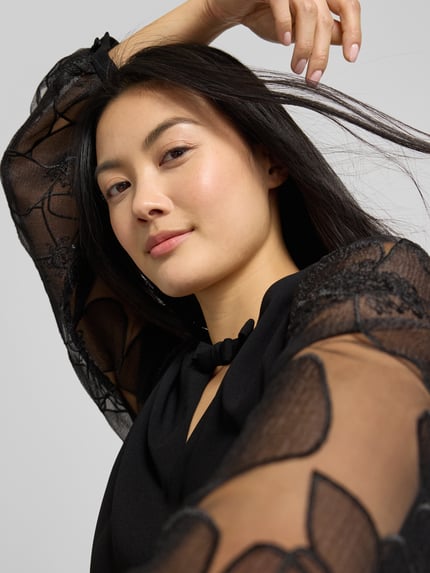Vrouwenmodel draagt Minikleid mit Ballonärmeln van Luisa Cerano in zwart, semitransparant met schlüsselloch-ausschnitt en strikdetail, detailopname bovenlichaam.
