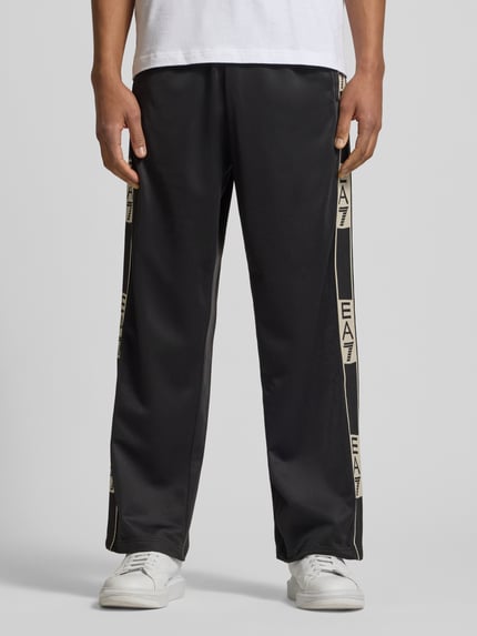 Männermodel trägt Trainingshose mit seitlichen Knopfleisten von EA7 Emporio Armani in Schwarz, Relaxed-Fit, Ganzkörperansicht von vorne.