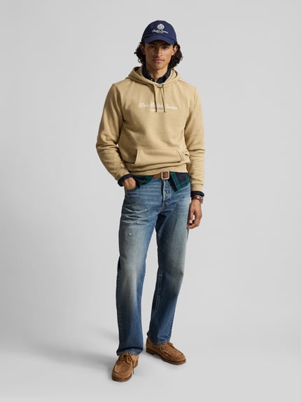 Männermodel trägt Relaxed Fit Hoodie aus Baumwoll-Mix von Polo Ralph Lauren in Beige, kombiniert mit blauer Jeans, Ganzkörperansicht von vorne. Herren Hoodie von Polo Ralph Lauren