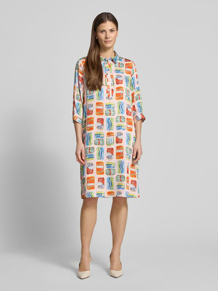 Modelka nosi knielanges Hemdblusenkleid aus Viskose im Allover-Look von Milano Italy, biały materiał z wielokolorowym wzorem, prosty krój, długość do kolan, kołnierzyk koszulowy, zapięcie na guziki, rękaw 3/4, długość tyłu w rozmiarze 36: 99 cm, szerokość w rozmiarze 36: 55 cm, długość rękawa w rozmiarze 36: 32 cm, widok całej sylwetki z przodu.