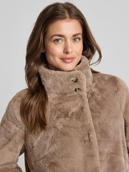 Frauenmodel trägt Jacke aus Kunstfell mit Eingrifftaschen von COMMA in Beige, weiche Struktur, Stehkragen, Nahaufnahme Oberkörper.