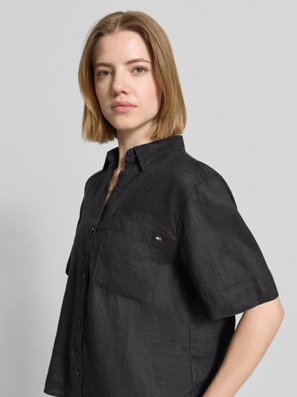 Kobiece model prezentuje Regular Fit Leinenbluse mit Logo-Stitching od Tommy Hilfiger, czarna, len, krótki rękaw, kieszeń na piersi, krój regular fit, kołnierzyk koszulowy, jednorzędowe zapięcie, detal logo, widok na górną część ciała z przodu