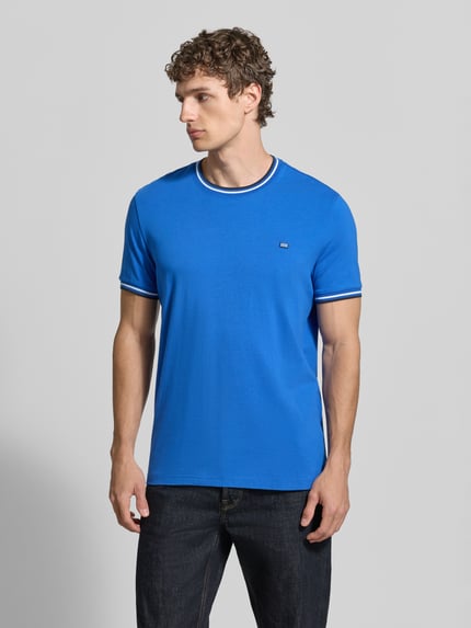 Męski model nosi T-Shirt mit Kontraststreifen und Rundhalsausschnitt od Christian Berg Men, bawełna z elastanem, krój regular fit, niebieski, widoczne kontrastowe paski na mankietach i dekolcie, długość tyłu w rozmiarze M 71 cm, widok całej sylwetki z przodu.