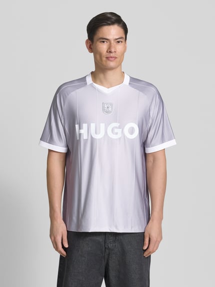 Męski model nosi Regular Fit T-Shirt mit Label Print model 'DWILD' od HUGO, wzór w paski, kolor szaro-biały, krótki rękaw, V-dekolt, detal logo, tekstura gładka, przód krój regular fit, całkowity widok z przodu.