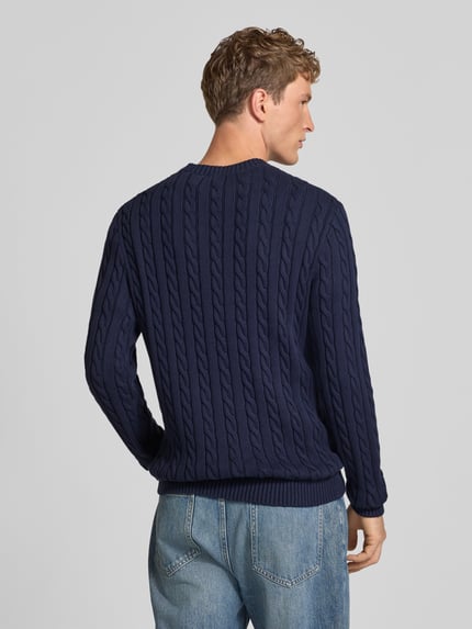 Męski model nosi Slim Fit Strickpullover aus reiner Baumwolle mit Zopfmuster von REVIEW, struktura ściegu warkoczowego, kolor granatowy, prążkowane wykończenia, długość tyłu w rozmiarze M: 70 cm, długość rękawów w rozmiarze M: 53 cm, widok całej sylwetki od tyłu.