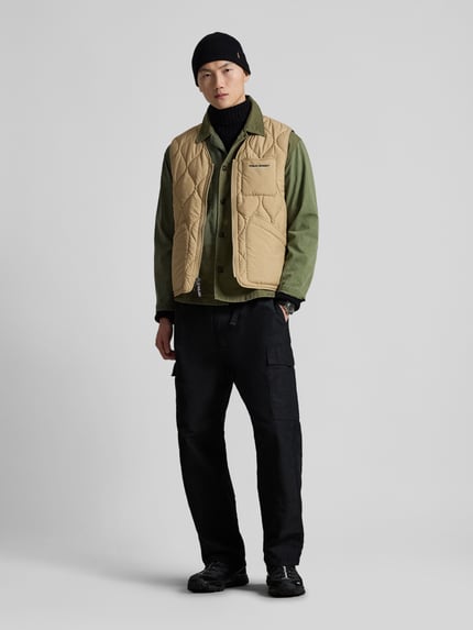 Männermodel trägt Regular Fit Steppweste mit Brusttasche von Polo Ralph Lauren in Beige, unifarben, V-Ausschnitt, Ganzkörperansicht von vorne.