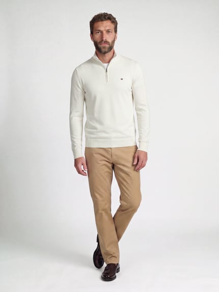 Męski model nosi Regular Fit Troyer aus reiner Baumwolle von Tommy Hilfiger, kremowy sweter, beżowe spodnie, brązowe buty, gładka tekstura, pełny widok z przodu.