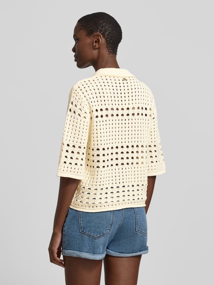 Vrouwenmodel draagt Regular Fit blouse in tricotlook met florale met siersteentjes van Liu Jo White, puur katoen, gaatjesmotief, platte kraag, halflange mouwen, ruglengte maat S 45 cm, mouwlengte maat S 34 cm, knoopsluiting, gebroken wit, denim short, geheel lichaamsaanzicht achteraan