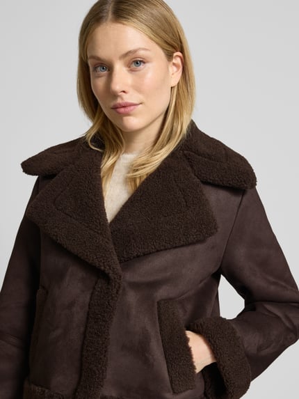 Kobieta prezentuje Regular Fit Kurtkę w imitacji skóry z podszewką z futerka model 'HARPER' od Vero Moda, efekt: jednolity kolor, kołnierz z połami, zapięcie na ukryte zatrzaski, długi rękaw, wpuszczane kieszenie, wykończenie futerkiem, całe zdjęcie od przodu, zbliżenie na górną część ciała.
