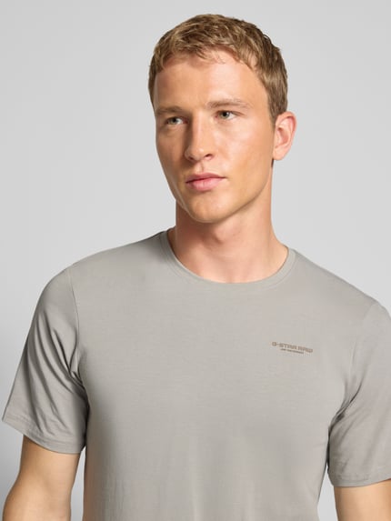 Męski model nosi Slim Fit T-Shirt mit Rundhalsausschnitt od G-Star Raw, szary, gładka tekstura, logo, bawełna z elastanem, zbliżenie na górną część ciała.