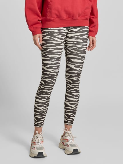 Kobiece model nosi legginsy z dodatkiem streczu od Juvia, wzór zebry, mieszanka materiałów, krój skinny fit, długość do kostek, szeroki elastyczny pas, długość wewnętrzna nogawki w rozmiarze S: 65 cm, szerokość w rozmiarze S: 34 cm, zdjęcie całej sylwetki z przodu.
