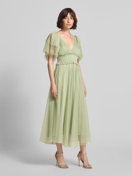 Frauenmodel trägt Midikleid mit Smok-Details und V-Ausschnitt von LACE & BEADS aus grünem Chiffon, A-Linie, Schmetterlingsärmel, Ganzkörperansicht von vorne. Rückenlänge bei Größe S: 35 cm, Brustweite bei Größe S: 29 cm.