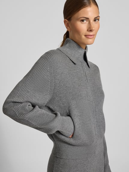 Frauenmodel trägt Regular Fit Strickjacke aus Schurwolle Modell 'FIBIONE' von DRYKORN in Grau, sichtbare Rippstruktur, Nahaufnahme Oberkörper.