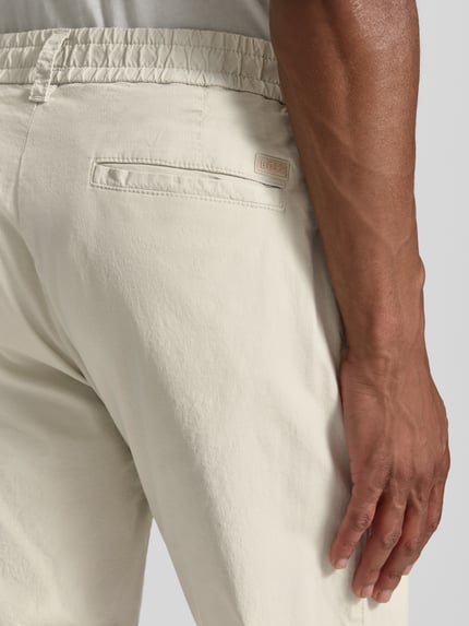 Männermodel trägt chinosy Tapered Chino aus Baumwoll-Mix Modell 'CHINO-TAPERED-DS-10' von BOSS Orange, efekt: jednolity kolor, krój tapered fit, szlufki na pasek, zapięcie na guzik i zamek błyskawiczny, tunel (do noszenia wewnątrz lub na zewnątrz), ściągacz: elastyczny, wpuszczane kieszenie, kieszeń z tyłu, materiał: mieszanka bawełny, detal: logo BOSS. Detailaufnahme Rückansicht.