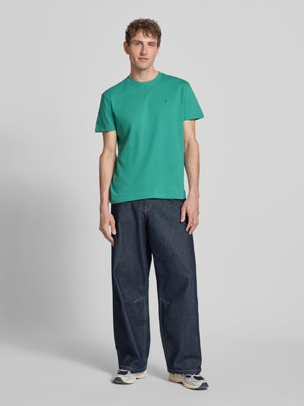 Männermodel trägt Regular Fit T-Shirt aus reiner Baumwolle von Tommy Jeans in Unifarbe, Rundhals, kurzarm, Logo-Stitching, Ganzkörperansicht von vorne.