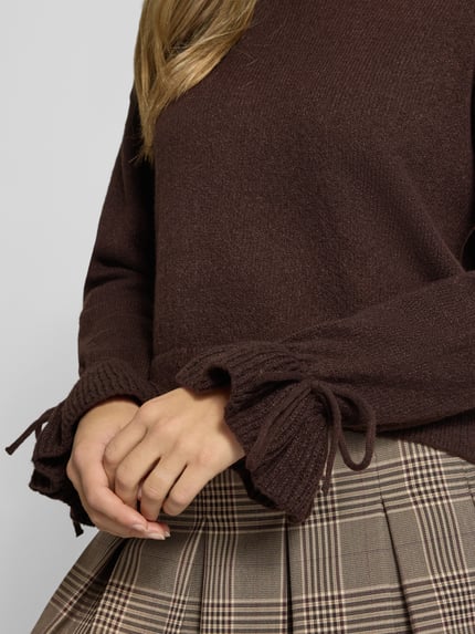 Frauenmodel trägt Pullover mit gerafften Ärmeln von PIECES in Braun, Strickoptik, Longline, Schleifen am Ärmel, Nahaufnahme Oberkörper.