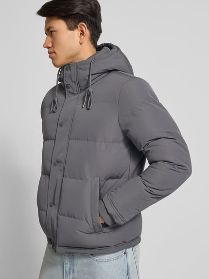 Männermodel trägt Steppjacke mit Eingrifftaschen Modell 'Everest' von Superdry in Grau, Regular-Fit, Kapuze, Label-Detail, Nahaufnahme Oberkörper