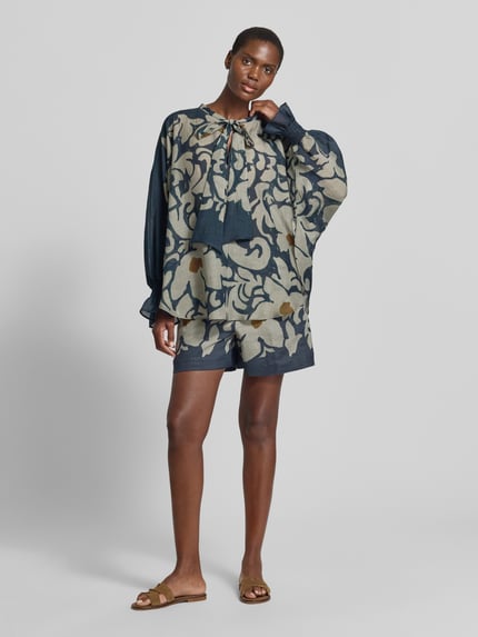 Modelka nosi Bluse mit Schluppe und Volantärmeln von Luisa Cerano, mieszanka lyocellu z dodatkiem ramii, krój loose fit, wycięcie w kształcie łezki z kokardą, rękawy z falbanami i marszczonym detalem, długość tyłu w rozmiarze 36: 74 cm, długość rękawów: 65 cm, wzór w odcieniach niebieskiego, beżowego i żółtego, tekstura lekko transparentna, cała sylwetka z przodu.