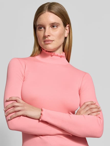 Frauenmodel trägt Langarmshirt mit Rollkragen-Rüschen von MARC CAIN in Uni-Rosa, Jersey mit Rollsaum, Nahaufnahme Oberkörper.