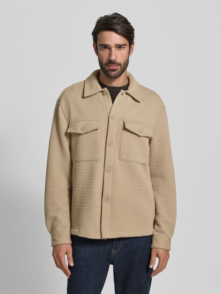Męski model nosi Hemdjacke mit Knopfleiste von MCNEAL z mieszanki materiałów z dodatkiem wiskozy, luźny krój, beż, kołnierzyk typu kent, kieszenie na piersi z zapięciem na guzik, długość tyłu 69 cm dla rozmiaru M, widok całej sylwetki z przodu.